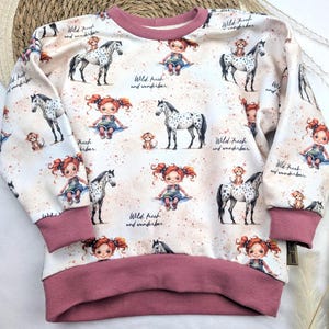 Puede incluir: Una sudadera blanca con ribete rosa presenta un patr&oacute;n repetido de una ni&ntilde;a montando un caballo con un mono en su espalda. El texto "Wild, frech and wunderbar" se repite en todo el dise&ntilde;o.