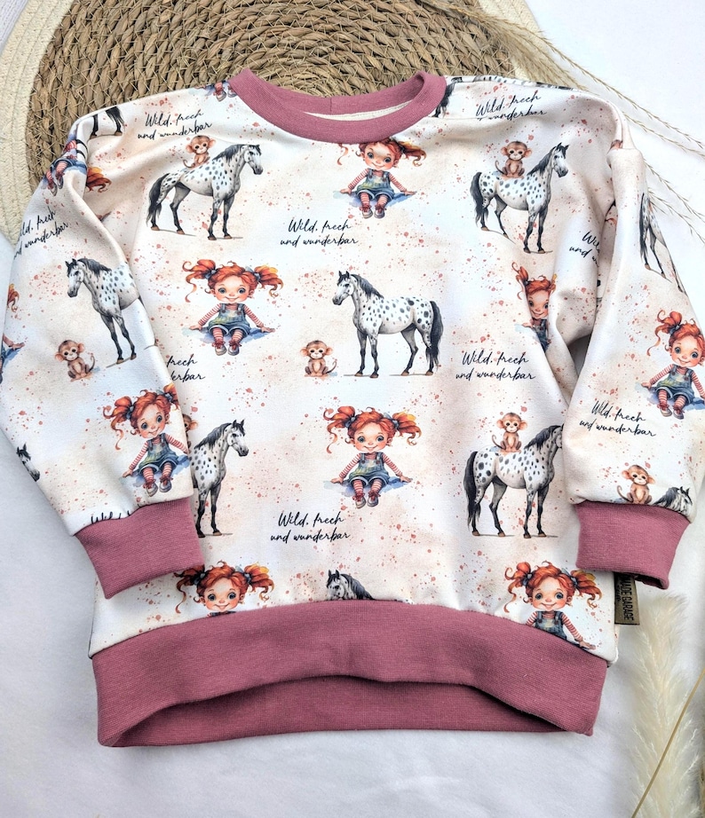Puede incluir: Una sudadera blanca con ribete rosa presenta un patr&oacute;n repetido de una ni&ntilde;a montando un caballo con un mono en su espalda. El texto "Wild, frech and wunderbar" se repite en todo el dise&ntilde;o.