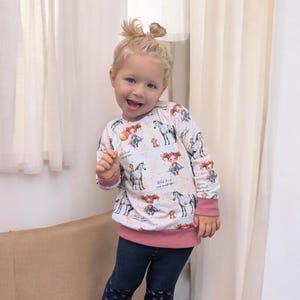Puede incluir: Una ni&ntilde;a peque&ntilde;a con una camisa de manga larga blanca con ribete rosa y estampado de caballos. La camisa tiene el texto "Wild heart and wanderlust" impreso. Lleva pantalones azules con corazones rosas y sostiene una piruleta.