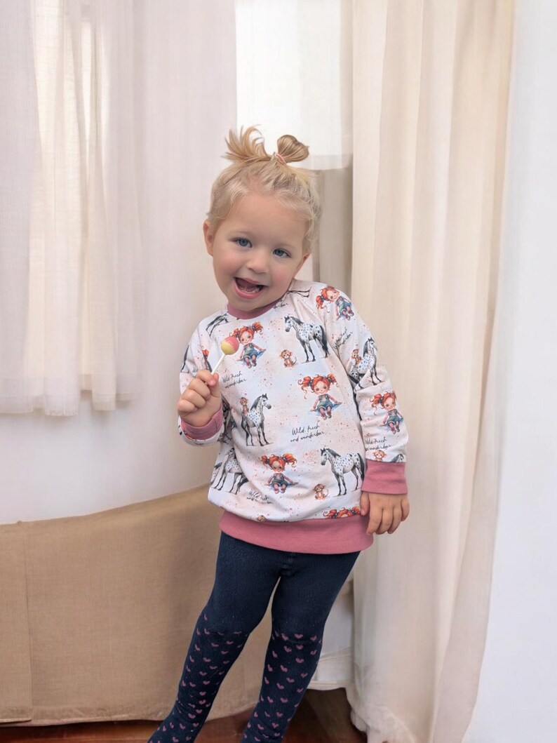 Puede incluir: Una ni&ntilde;a peque&ntilde;a con una camisa de manga larga blanca con ribete rosa y estampado de caballos. La camisa tiene el texto "Wild heart and wanderlust" impreso. Lleva pantalones azules con corazones rosas y sostiene una piruleta.