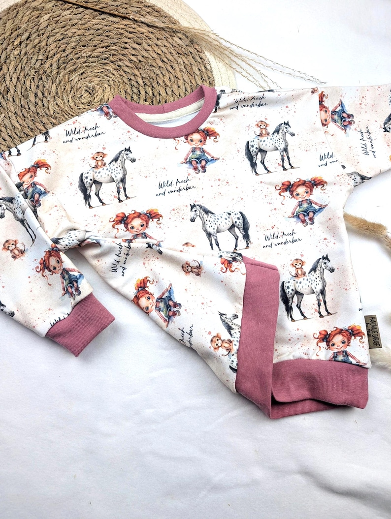 Puede incluir: Una sudadera blanca de manga larga con ribetes rosas. La sudadera tiene un patr&oacute;n repetido de una ni&ntilde;a con cabello rojo montando un caballo manchado. El texto "Wild. Frech und wunderbar" se repite en todo el patr&oacute;n.