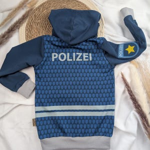 Könnte beinhalten: Ein blauer Kapuzenpullover mit dem weißen Schriftzug "POLIZEI" auf der Rückseite und einem Muster aus blauen und weißen Schilden. Der rechte Ärmel hat einen blauen Aufnäher mit einem gelben Stern und dem Wort "POLIZEI" darauf.