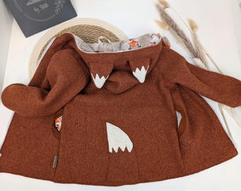 May include: A rust-colored hooded jacket made of boiled wool. The hood features two pointed ears with white tips, and the back of the jacket displays a white triangular tail detail. The interior lining is patterned with small fox illustrations.