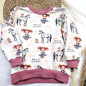 Puede incluir: Una sudadera blanca con ribete rosa presenta un patr&oacute;n repetido de una ni&ntilde;a montando un caballo manchado, un mono y el texto "Wild. frech and wunderbar".