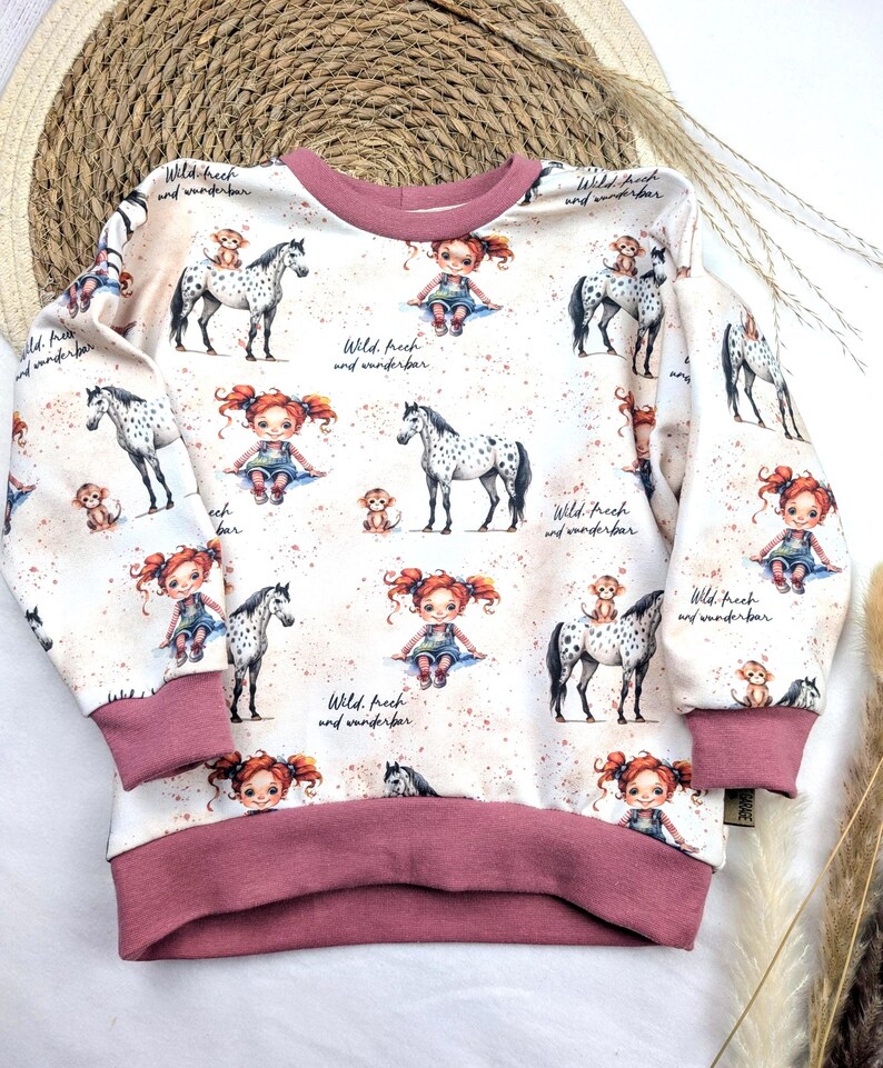 Puede incluir: Una sudadera blanca con ribete rosa presenta un patr&oacute;n repetido de una ni&ntilde;a montando un caballo manchado, un mono y el texto "Wild. frech and wunderbar".