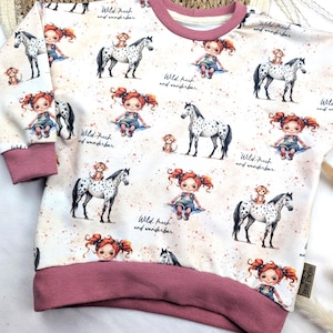 Puede incluir: Una sudadera blanca con ribete rosa presenta un patr&oacute;n repetido de una ni&ntilde;a con cabello rojo montando un caballo manchado. El texto "Wild. frech and wunderbar" se repite en todo el patr&oacute;n.