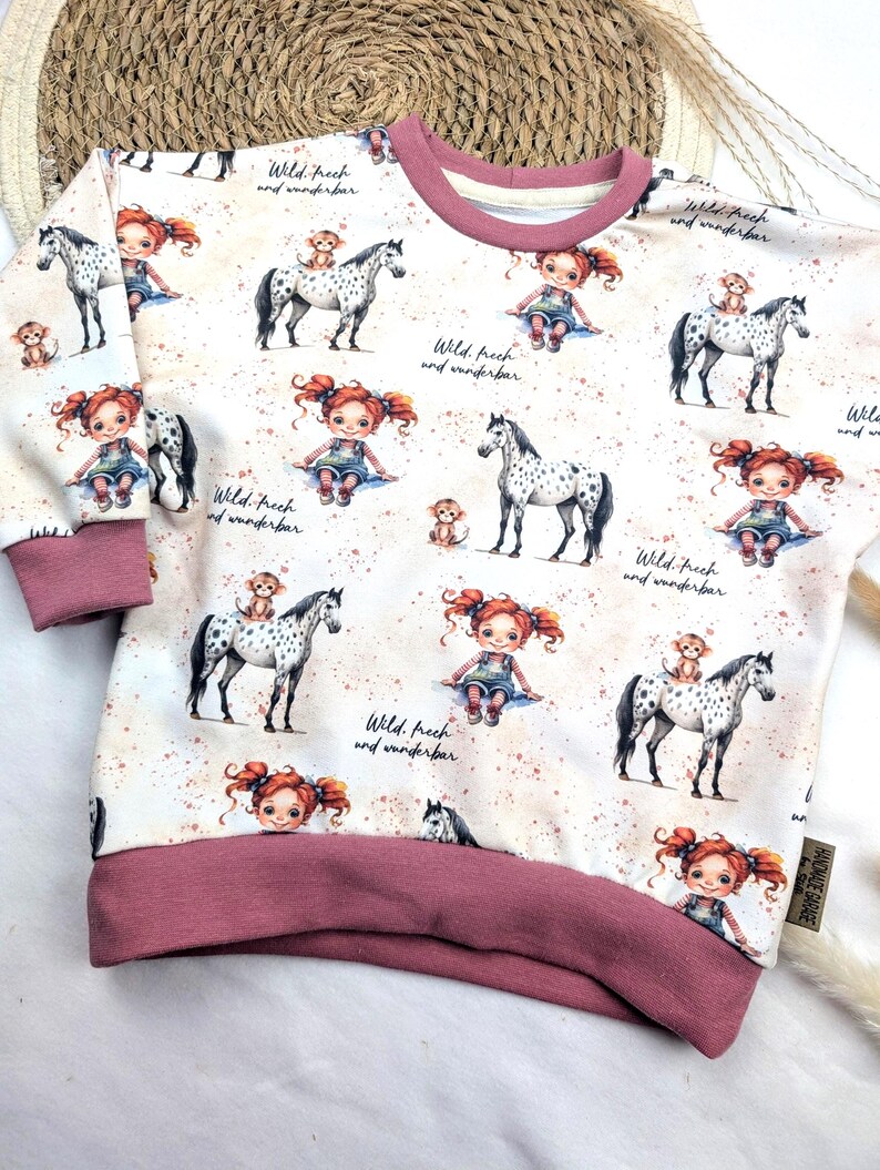 Puede incluir: Una sudadera blanca con ribete rosa presenta un patr&oacute;n repetido de una ni&ntilde;a con cabello rojo montando un caballo manchado. El texto "Wild. frech and wunderbar" se repite en todo el patr&oacute;n.