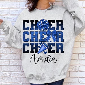 May include: A gray sweatshirt with the words "Cheer Cheer Cheer" in blue glitter lettering. A blue and white pom pom and a blue megaphone are above the text. The name "Amelia" is printed below the text.