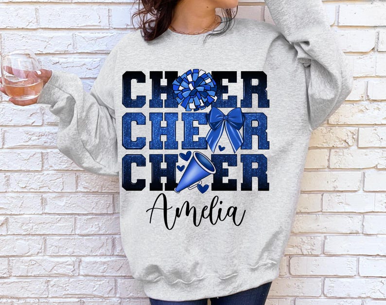 May include: A gray sweatshirt with the words "Cheer Cheer Cheer" in blue glitter lettering. A blue and white pom pom and a blue megaphone are above the text. The name "Amelia" is printed below the text.