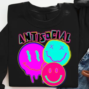 May include: Black sweatshirt with the word "ANTISOCIAL" in neon lettering above three smiley face graphics. The smiley faces are in pink, blue, and pink with neon yellow and blue accents. The sweatshirt is folded.