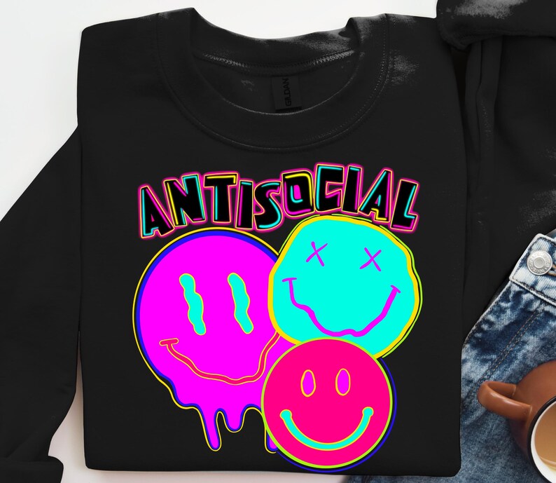 May include: Black sweatshirt with the word "ANTISOCIAL" in neon lettering above three smiley face graphics. The smiley faces are in pink, blue, and pink with neon yellow and blue accents. The sweatshirt is folded.