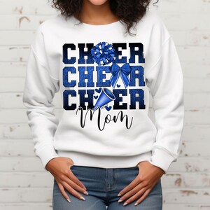 May include: White sweatshirt with blue glitter text that reads "Cheer Cheer Cheer Mom". The text is layered with a blue pom pom, a blue bow, and a blue megaphone.