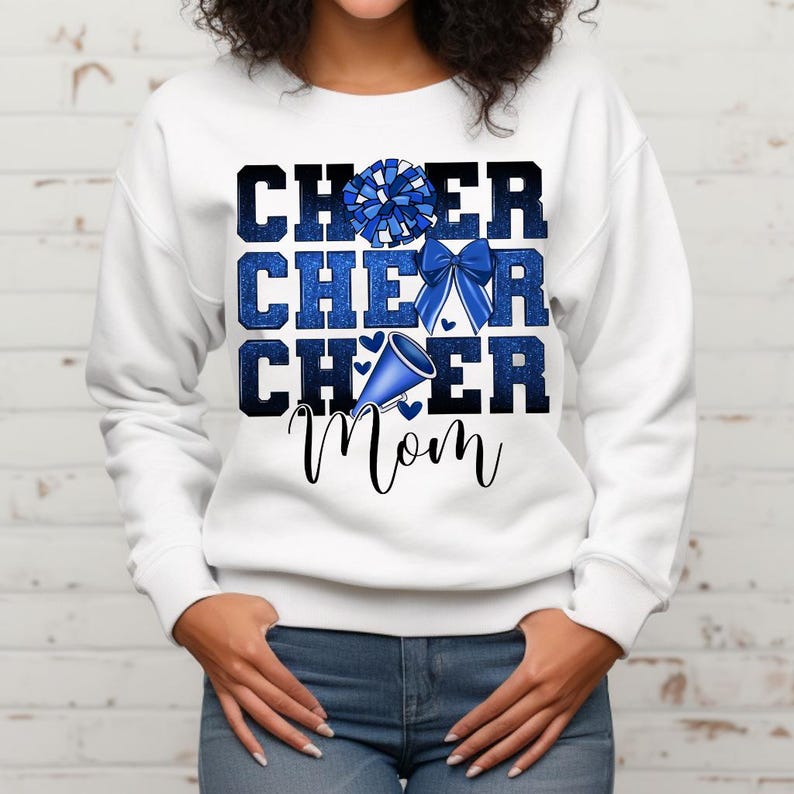 May include: White sweatshirt with blue glitter text that reads "Cheer Cheer Cheer Mom". The text is layered with a blue pom pom, a blue bow, and a blue megaphone.