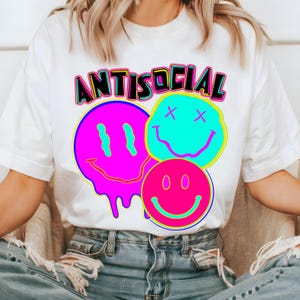 May include: White t-shirt featuring the word "ANTISOCIAL" in black, outlined in yellow and pink, above three smiley face graphics. The smiley faces are in pink, blue, and pink with black details.