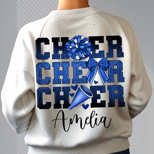May include: White sweatshirt with blue glitter text that reads "Cheer Cheer Cheer" with a blue pom pom and a blue megaphone. The name "Amelia" is written in black below the text.