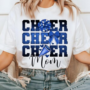 May include: White t-shirt with blue glitter text that reads "Cheer Cheer Cheer Mom" with a blue and white pom pom, a blue bow, and a blue megaphone.
