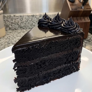 Könnte beinhalten: Ein Stück dunkler Schokoladenkuchen mit drei Schichten aus saftigem Biskuit, die durch dünne Schichten Schokoladencreme getrennt sind, bedeckt mit einer glatten Schokoladenglasur und drei dekorativen, aufgespritzten Cremetupfern.