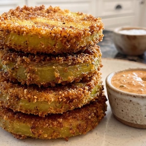 May include: A stack of four breaded and fried green tomato slices with a golden-brown, crispy exterior. Beside the stack sits a small, speckled ceramic bowl filled with a creamy, orange-colored dipping sauce and a second small bowl of white sauce.