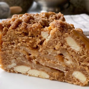 May include: A thick slice of golden-brown cake featuring a crumbly streusel topping, visible chunks of white chocolate, and layers of sliced apples. A drizzle of caramel sauce is visible on the top edge of the dense, textured baked dessert.