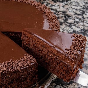 Può includere: Una ricca torta al cioccolato con una copertura di ganache liscia e lucida e un bordo di piccole scaglie di cioccolato. Una singola fetta triangolare viene sollevata dalla torta principale con una spatola in metallo.