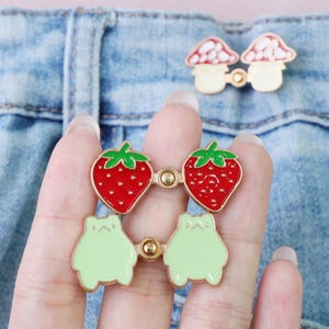 May include: Enamel pins with a strawberry, frog, and mushroom design. The strawberry pins are red with green leaves, the frog pins are light green with simple faces. The mushroom pins have a cream base and pink and white tops.