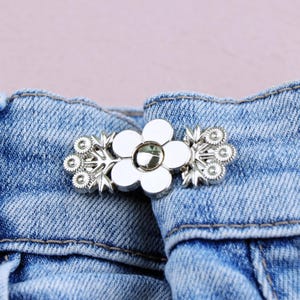 May include: A silver-toned flower-shaped button clasp with a central gem, attached to blue denim fabric. The clasp features a floral design with a central flower and decorative elements on each side, providing a decorative closure.