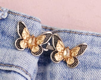 May include: Two butterfly-shaped jean buttons with gold-colored wings and silver-colored borders are attached to a blue denim garment. The butterflies have a gemstone in the center. The background is a light pink color.