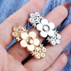 May include: Close-up of two flower-shaped hair accessories. One is gold-toned, the other is silver-toned. Each has a central flower with surrounding smaller flower accents. The accessories are on a black band.