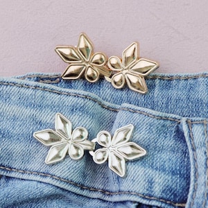 May include: Close-up of two sets of decorative metal clasps, each featuring a floral design with five petals. The clasps are gold and silver, and are attached to a blue denim fabric.