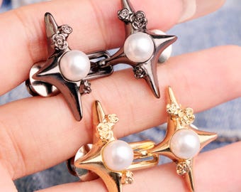 May include: Four star-shaped cufflinks. Two are gunmetal gray with pearl centers and floral accents. The other two are gold-toned with pearl centers and floral accents. The cufflinks are displayed on a light blue denim background.