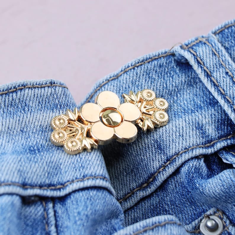 May include: A gold-colored metal flower-shaped brooch with a central flower and floral accents. The brooch is attached to a blue denim garment, showcasing its decorative design and potential use as a fashion accessory.