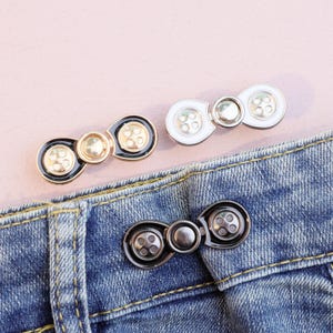 May include: Three sets of decorative jean button extenders in gold, black, white, and silver. The extenders are attached to a blue denim fabric. The extenders have a button design.