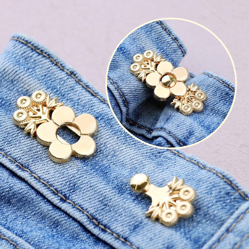 May include: Gold-colored flower-shaped jean buttons. The buttons have a floral design with a central flower and smaller floral accents. The buttons are attached to a blue denim fabric.