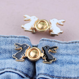 Peut inclure: Deux ensembles de fermoirs décoratifs en forme de lapin. Un ensemble présente des lapins blancs avec des accents dorés, et l'autre des lapins noirs avec des accents dorés. Les fermoirs sont fixés à un tissu en denim, montrant leur utilisation comme accessoire vestimentaire.