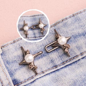 May include: Set of two star-shaped metal jean buttons with a pearl center and small rose accents. The buttons are a dark gunmetal color and are attached to a light blue denim fabric. The buttons are designed to replace traditional jean buttons.
