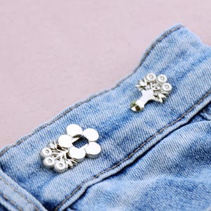May include: Close-up of two silver-colored flower-shaped brooches on a blue denim fabric. One brooch is a simple flower design, while the other features a bouquet of flowers. The brooches are attached to the denim.
