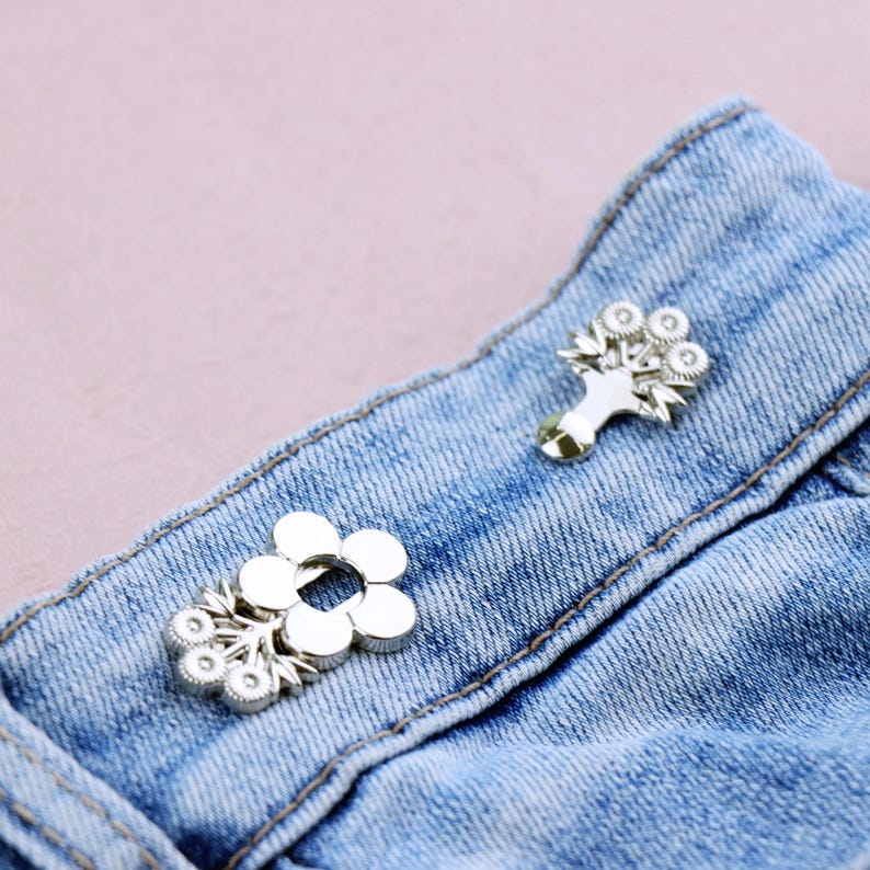 May include: Close-up of two silver-colored flower-shaped brooches on a blue denim fabric. One brooch is a simple flower design, while the other features a bouquet of flowers. The brooches are attached to the denim.
