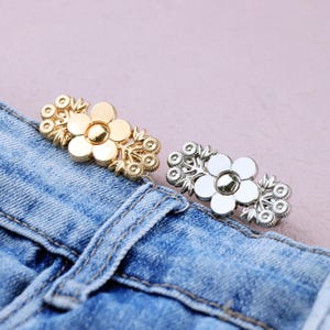 May include: Two flower-shaped decorative buttons, one gold and one silver, are displayed on a blue denim fabric. The buttons have a floral design with a central gem and surrounding petal details. The buttons are likely used for clothing or accessories.
