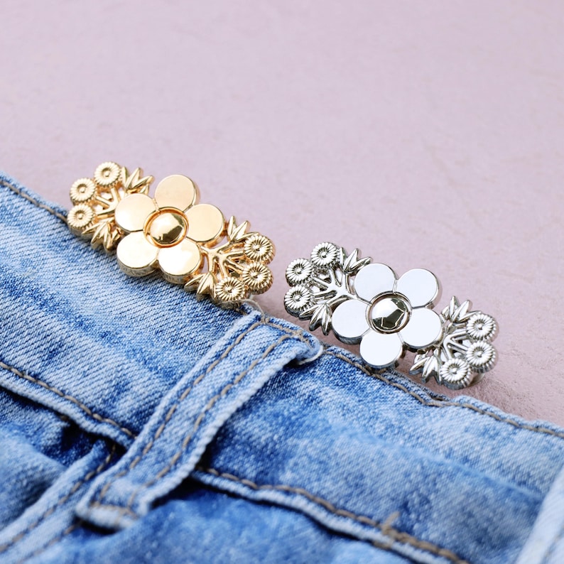 May include: Two flower-shaped decorative buttons, one gold and one silver, are displayed on a blue denim fabric. The buttons have a floral design with a central gem and surrounding petal details. The buttons are likely used for clothing or accessories.