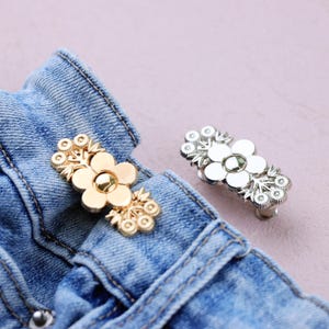 Könnte beinhalten: Zwei dekorative Jeansknöpfe in Blumenform, einer goldfarben und einer silberfarben, werden auf einem Jeanskleidungsstück gezeigt. Die Knöpfe haben ein Blumendesign mit einer zentralen Blume und dekorativen Akzenten.