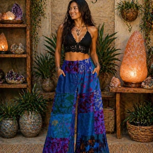 Könnte beinhalten: Eine Frau trägt ein schwarzes Häkeltop und eine Patchwork-Hose mit weitem Bein in Blau, Lila und Grün mit floralem Muster. Im Hintergrund steht der Text "Zamla & Gems" und dekorative Regale.