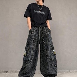 Könnte beinhalten: Ein schwarzes kurzärmeliges T-Shirt mit grauem Grafikdruck auf der Brust, kombiniert mit einer weit geschnittenen, säuregewaschenen schwarzen Jeans-Cargohose mit mehreren Taschen, Kordelzug in der Taille und kleinen gestickten Pilzaufnähern.