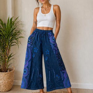 Peut inclure: Pantalon large en jean avec broderies florales et mandalas bleus et violets. Le pantalon a une taille à cordon et est porté avec un débardeur blanc côtelé. La personne porte des sandales marron.