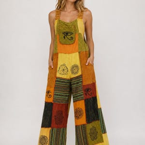 May include: A sleeveless jumpsuit featuring a patchwork design in shades of yellow, orange, green, and black. The garment includes a front pocket, wide legs, and various printed symbols such as the Eye of Horus, suns, and Hamsa hands.