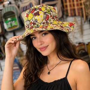 Peut inclure: Un chapeau bob orné d'un motif floral et fruité multicolore aux tons rouge, jaune et vert sur un fond clair. Ce chapeau présente un bord large incliné vers le bas et une calotte arrondie, fabriqué dans un tissu à la texture apparente.