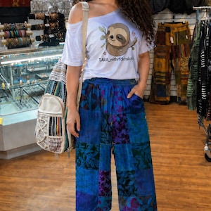 Può includere: Una donna indossa una maglietta bianca con spalle scoperte con una grafica di bradipo e pantaloni patchwork blu. I pantaloni sono cropped e hanno una vita con coulisse. Indossa scarpe da ginnastica bianche e porta uno zaino.