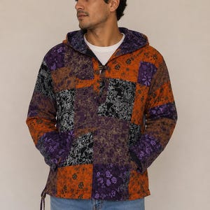 Puede incluir: Una sudadera con capucha y manga larga que presenta un diseño de patchwork con paneles de tela en naranja, morado y negro. La prenda incluye una camiseta interior blanca, un cierre de palanca en el cuello, dos bolsillos y un cordón ajustable.