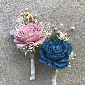 May include: Two boutonnieres with wooden rose flowers. One is dusty rose, the other is blue. Both have white ribbon and twine wrapped stems, with small white and yellow accent flowers.