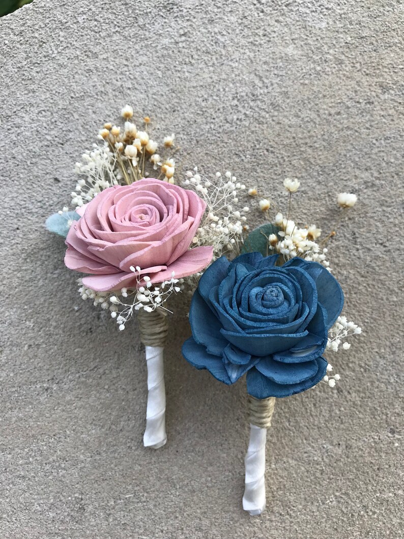 May include: Two boutonnieres with wooden rose flowers. One is dusty rose, the other is blue. Both have white ribbon and twine wrapped stems, with small white and yellow accent flowers.