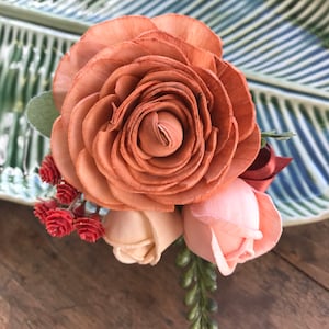 May include: A handcrafted floral arrangement featuring a large, light brown rose as the focal point, complemented by smaller peach and cream-colored roses. Green and red accents add contrast to the arrangement, which is set against a green and white leaf-patterned background.